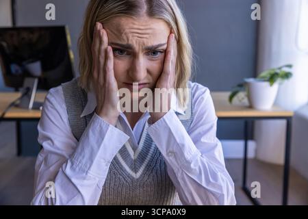 Donna che indossa un gilet grigio su una camicia bianca che tiene le aste alla scrivania dell'ufficio accanto al monitor Foto Stock
