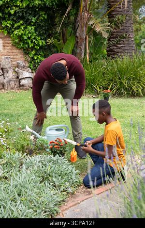 Padre e figlio afroamericani che usano la cazzuola e le lattine d'acqua nel giardino sul retro Foto Stock