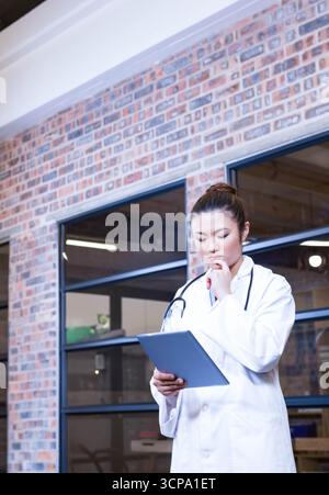 Medico asiatico che indossa lo stetoscopio usando tavoletta, che riposa il mento a portata di mano nel corridoio della clinica di mattoni Foto Stock