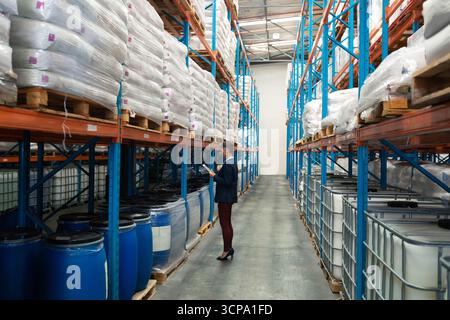 Donna anziana in blazer che ispeziona l'inventario su tablet nel corridoio del magazzino con sacchi e barili Foto Stock