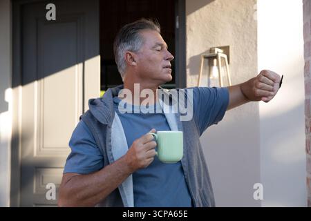 Uomo anziano che tiene una tazza pallida sul portico anteriore, controllando l'orologio sotto la luce del sole mattutino Foto Stock