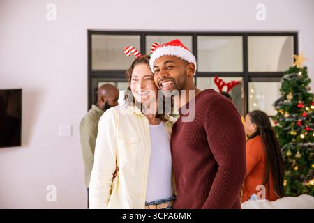 Coppia variegata che indossa il cappello di babbo natale e la fascia di canna da zucchero che si abbraccia a casa accanto all'albero di natale Foto Stock