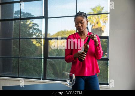 Donna afroamericana che apre la bottiglia di vino al tavolo vicino alla finestra a griglia con bicchiere vuoto, copia spazio Foto Stock