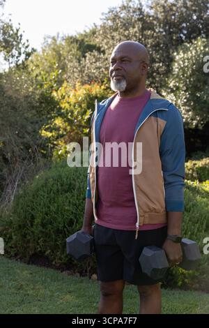 Uomo afroamericano senior che solleva manubri neri in giardino indossando giacca e smartwatch Foto Stock
