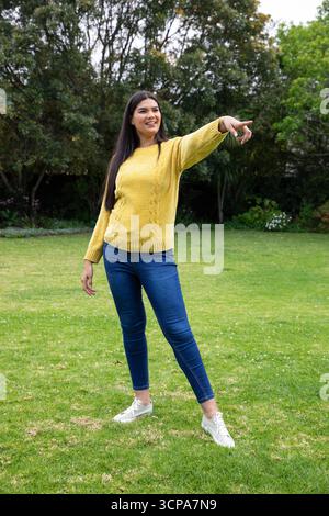 Donna asiatica in piedi nel cortile con maglione giallo e jeans che puntano ad alberi, arbusti Foto Stock