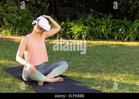 Donna asiatica di mezza età seduta su un tappetino da yoga nel cortile posteriore con visore VR e collo allungato Foto Stock