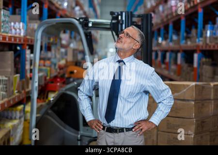 Uomo anziano che indossa camicia e cravatta che ispeziona gli scaffali con carrello elevatore, pallet avvolti in magazzino Foto Stock