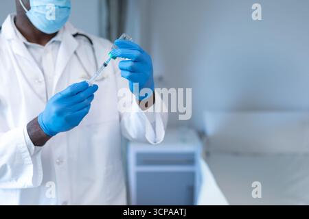 Medico afroamericano di sesso maschile che aspira il farmaco dal flaconcino alla siringa in clinica, copia spazio Foto Stock