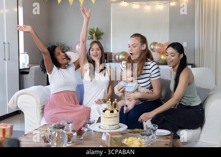 Diverse amiche che tengono in braccio il bambino e festeggiano sul divano a casa con una torta di compleanno e regali Foto Stock