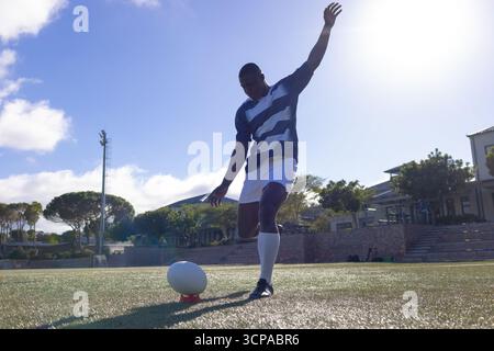 Uomo afro-americano che indossa una maglia a righe che calcia la palla da rugby su un tee rosso su un campo da rugby in erba Foto Stock