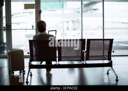 Donna d'affari di mezza età seduta sulla panchina al cancello dell'aeroporto con un tablet con valigia, spazio per copiare Foto Stock