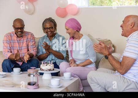 Diversi amici anziani seduti sul divano in casa per festeggiare il compleanno con candele per torte e palloncini Foto Stock