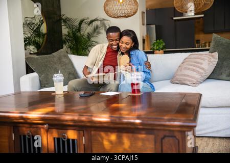 Coppia afroamericana che abbraccia e condivide il takeout nel salotto di casa accanto al tavolino da caffè con tazze Foto Stock