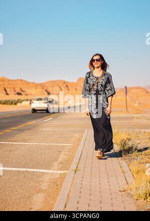 Giovane donna con lungo abito nero che cammina sul marciapiede vicino alla strada - paesaggio roccioso del deserto, tipico di Alula, sullo sfondo Foto Stock