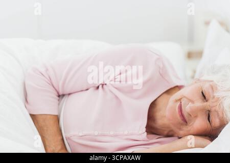 Donna anziana sdraiata sul letto con pigiama rosa con cuscino bianco e piumone Foto Stock