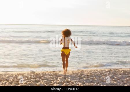 Donna afroamericana che cammina su sabbia bagnata nelle onde poco profonde dell'oceano sulla spiaggia indossando un bikini giallo Foto Stock