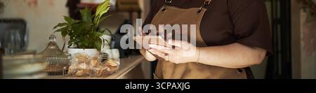 Giovane donna caucasica adulta in piedi dietro il bancone con tablet digitale, mani visibili vicino a prodotti da forno e piante in vaso, grembiule, lavoro in un bar o in un panificio Foto Stock