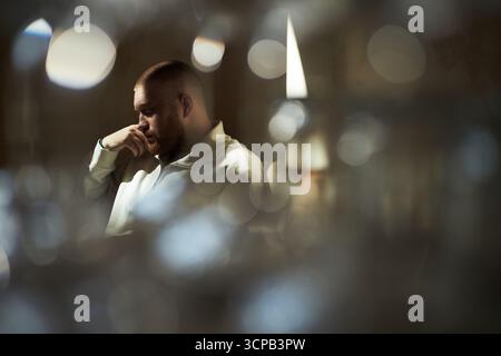 Giovane uomo adulto caucasico seduto con la mano appoggiata sul viso, che appare profondo nel pensiero, capelli corti e barba rifinita visibili, elementi in primo piano sfocati che incorniciano il soggetto Foto Stock