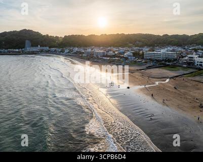 Vista aerea delle tranquille sabbie baciate dal sole della spiaggia che incontrano le onde ondulate dell'oceano, con la città costiera annidata sotto le colline verdeggianti, Kamakura, Kanagawa, Giappone. Foto Stock