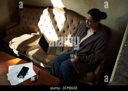 Giovane donna caucasica seduto sul divano con lo smartphone e in mano un bicchiere d'acqua, aprire il computer portatile e i documenti con le carte e lo smartphone sul tavolo davanti Foto Stock