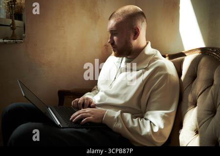 Giovane adulto caucasico seduto sul divano con il laptop, focalizzato sullo schermo con le mani sulla tastiera, luce naturale che illumina il viso, barba corta e testa rasata visibile Foto Stock