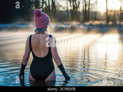 Una donna, vista da dietro, si ferma durante una nuotata di acqua fredda al mattino presto, godendosi il tranquillo e nebbioso lago e la luce dorata. Foto Stock
