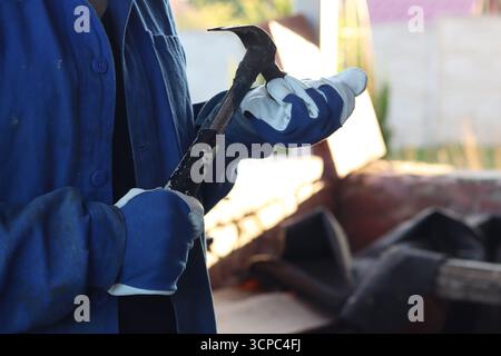 Donna in abiti da lavoro con un martello da chiodatrice, mani da vicino. Donna in guanti da lavoro con un utensile manuale in mano. Persona irriconoscibile con un martello Foto Stock