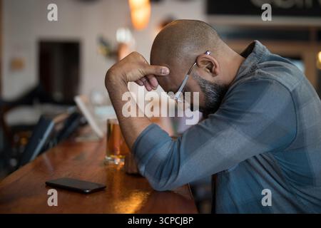 Uomo indiano anziano seduto al banco di legno nel pub con smartphone e pinta mezza piena, spazio fotocopie Foto Stock