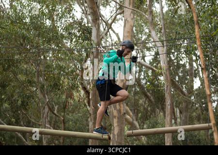 Donna adulta che attraversa travi di legno su corde tra tronchi d'albero con imbracatura e casco Foto Stock