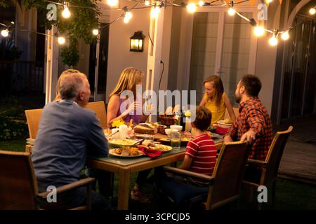 Famiglia di più generazioni che chiacchiera intorno al tavolo da pranzo in vimini sul patio sul cortile posteriore sotto le luci a corde Foto Stock