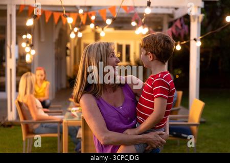 Riunione di famiglia multigenerazionale a tavola sotto pergola con luci a corda e accatastamento Foto Stock