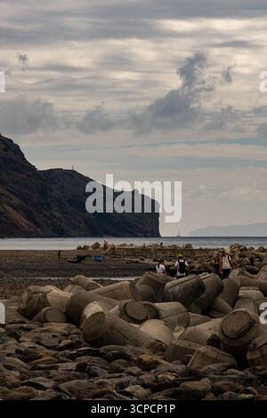 I visitatori camminano lungo una spiaggia rocciosa e un frangiflutti in cemento, con piccole barche sbarcate e una grande parete rocciosa che si innalza sullo sfondo sotto una colomba Foto Stock