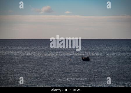 Una piccola barca da pesca tradizionale, probabilmente gestita da pescatori locali, galleggia tranquillamente in mare aperto sotto un cielo nebbioso, impegnata in fis o in preparazione per la fis Foto Stock