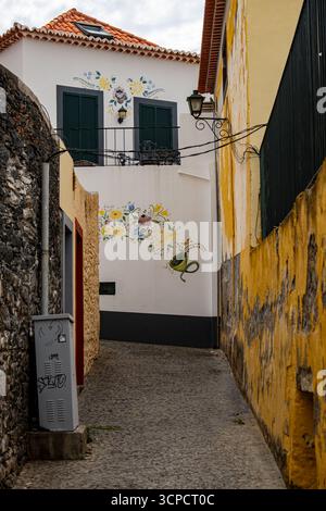 Una stretta strada acciottolata in una cittadina europea, che presenta un edificio bianco con tetto in terracotta, un balcone con persiane verdi scure e persiane Foto Stock