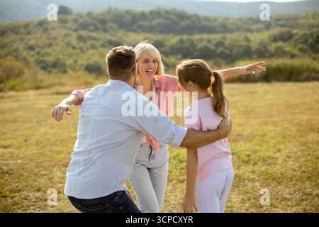 In un pittoresco scenario di campagna, un uomo e una ragazza abbracciano una donna con gioia sui loro volti. Colline lussureggianti incorniciano lo sfondo sotto un cielo blu chiaro Foto Stock