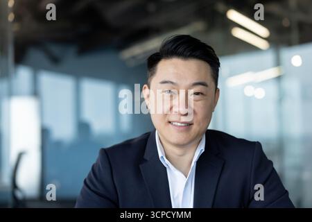 Primo piano ritratto di un giovane uomo d'affari asiatico seduto alla scrivania in ufficio e sorridente alla macchina fotografica. Foto Stock