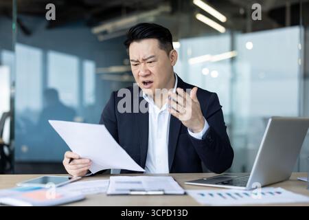 Il giovane asiatico arrabbiato ha ricevuto una lettera e cattive notizie, seduto deluso nell'ufficio, leggendo e tenendo in mano documenti. Foto Stock