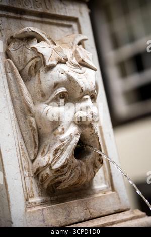 Un primo piano di una fontana decorativa in pietra presenta una grottesca intagliata o un volto simile a satiro con una bocca aperta, da cui un flusso d'acqua continuo Foto Stock