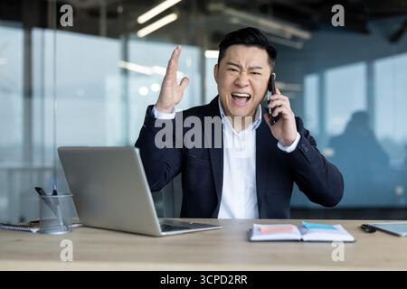 Giovane asiatico arrabbiato seduto a una scrivania in ufficio, a parlare e urlare al telefono. Foto Stock