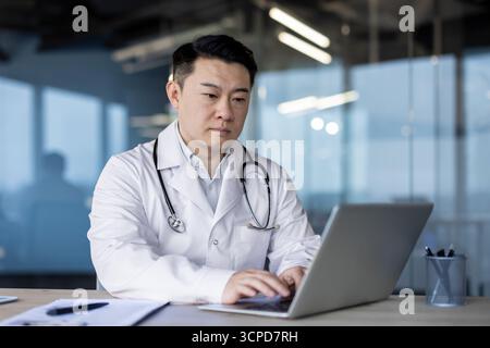 Serio giovane asiatico maschio medico in cappotto bianco che lavora intensamente al laptop. Foto Stock