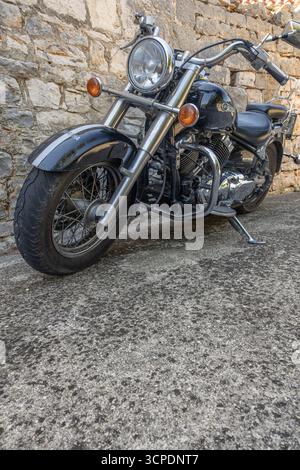 Una classica motocicletta cruiser nera parcheggiata accanto a una rustica parete in pietra, con accenti cromati e stile vintage. Semplice, robusto e senza tempo: Perfetto per te Foto Stock