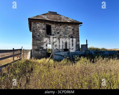 Vista ravvicinata di una storica fattoria in pietra abbandonata circondata da alte praterie e da rustiche recinzioni in legno sotto un cielo azzurro. Foto Stock