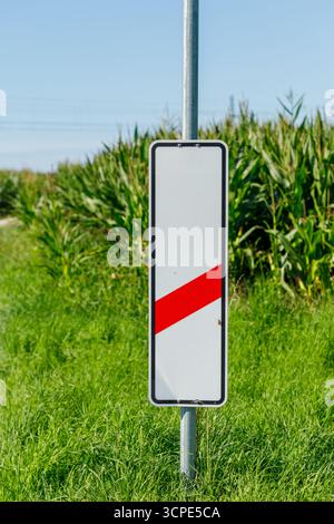 Un segnale di attraversamento ferroviario con una striscia rossa si trova in un campo erboso, sostenuto da gambi di mais sotto un cielo azzurro. Foto Stock