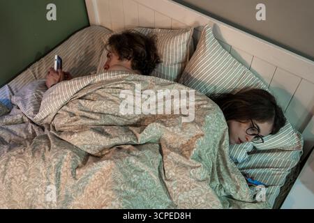 La coppia sdraiata a letto si ignora a vicenda mentre utilizza gli smartphone, illustrando dipendenza digitale, problemi di relazione, disconnessione emotiva e problemi di stile di vita moderno. Foto Stock