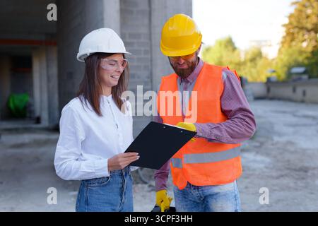 Architetto e caposquadra che rivede i progetti su una clipboard, discutendo i progressi e i piani nel cantiere Foto Stock