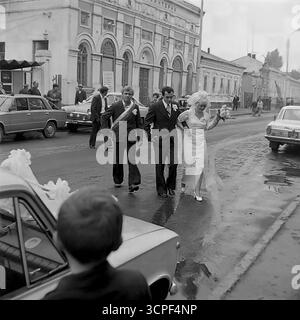 Una coppia appena sposata cammina in una processione nuziale lungo una strada bagnata a Sloviansk, RSS Ucraina, nel 1976. Questa serie monocromatica di archivio cattura un momento sincero di un matrimonio di epoca sovietica, con una classica GAZ-21 Volga come auto da sposa e veicoli VAZ-2101 che accompagnano. La scena, sullo sfondo del centro storico della città in Lenin Street, riflette le tradizioni sociali, la moda degli anni '1970 e l'atmosfera autentica di una città di provincia Foto Stock