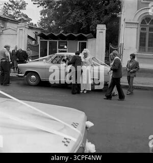 Una coppia appena sposata cammina in una processione nuziale lungo una strada bagnata a Sloviansk, RSS Ucraina, nel 1976. Questa serie monocromatica di archivio cattura un momento sincero di un matrimonio di epoca sovietica, con una classica GAZ-21 Volga come auto da sposa e veicoli VAZ-2101 che accompagnano. La scena, sullo sfondo del centro storico della città in Lenin Street, riflette le tradizioni sociali, la moda degli anni '1970 e l'atmosfera autentica di una città di provincia Foto Stock