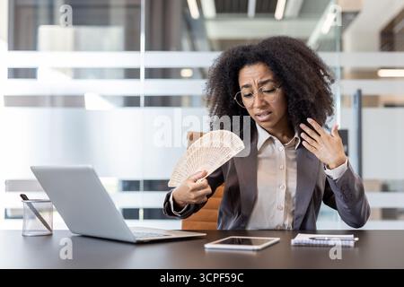 Giovane donna d'affari afroamericana seduta alla sua scrivania soffrendo del caldo, agitando un ventilatore in faccia. Foto Stock