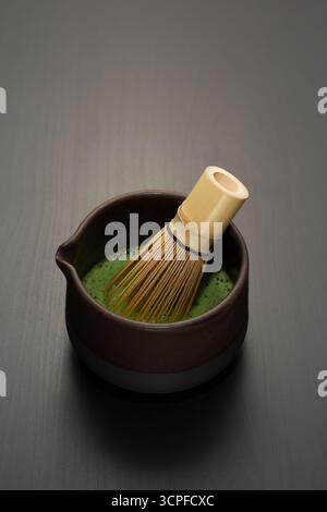 Una frusta chasen di bambù si appoggia in un chawan artigianale di vibranti matcha verdi, la sua superficie schiumosa che contrasta con i toni caldi e terrosi della ciotola Foto Stock