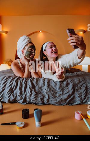 Due allegre donne si stanno facendo un selfie con uno smartphone mentre sono sdraiate sul letto e indossano maschere cosmetiche per il viso, per godersi una rilassante giornata in spa a casa Foto Stock